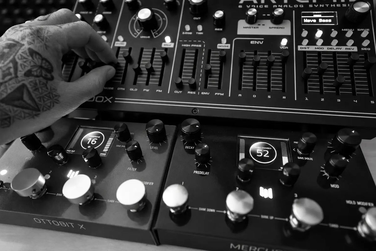 Close-up of a tattooed hand adjusting an Artemis synthesizer above two Meris effects pedals, in black and white.
